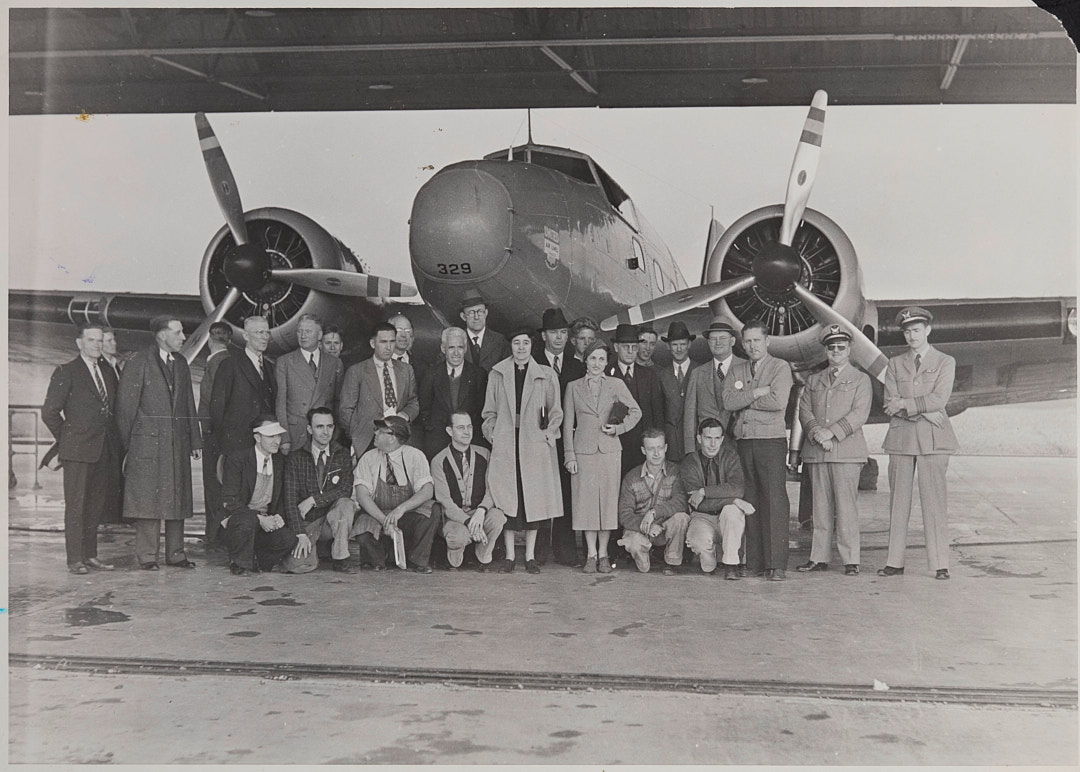 Black-and-white photograph of a large group of adults standing in front of an airplane inside a hangar.
