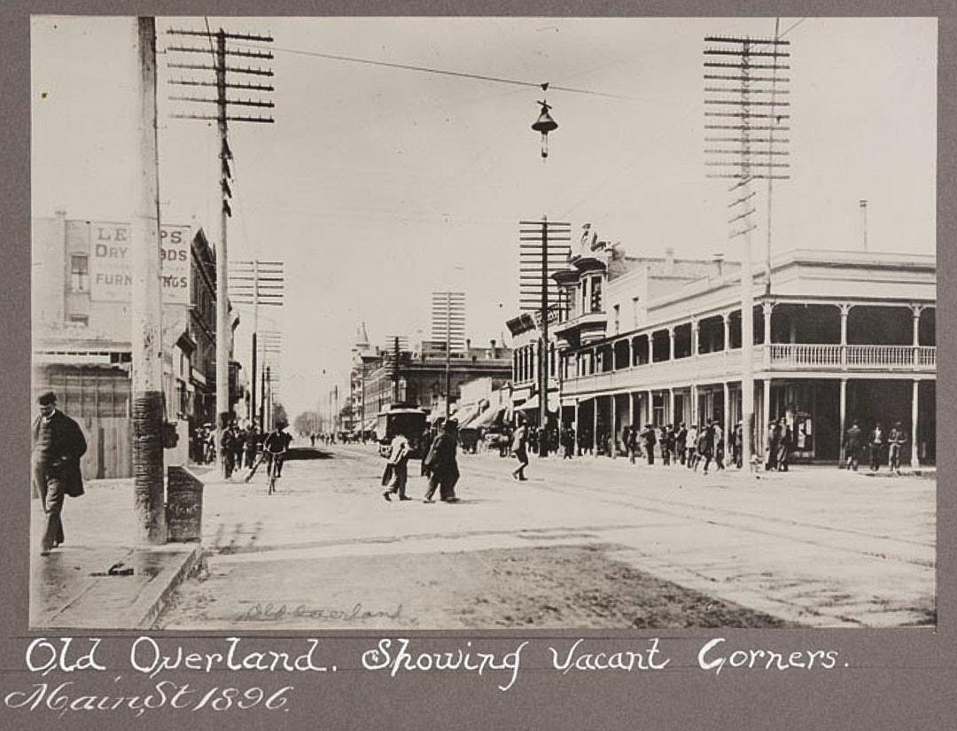Black-and-white photograph of a city street lined with buildings, and people walking and riding bicycles along sidewalks and on a dirt road. Large utility poles are visible on both sides of the street. 