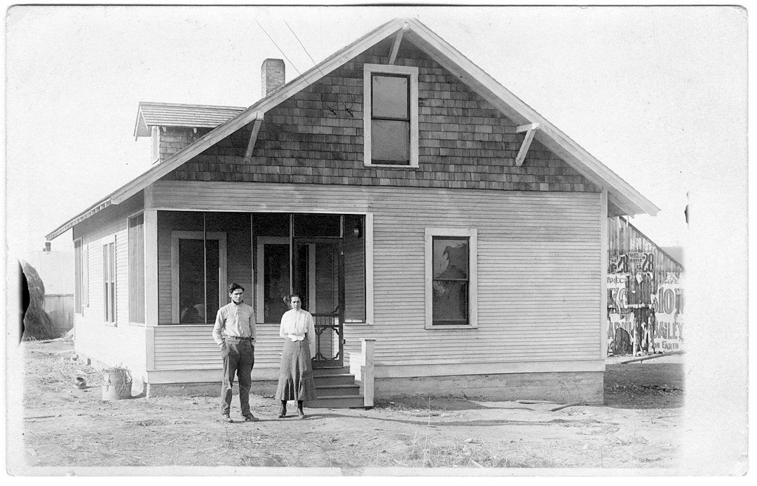 A woman and man stand in front of a two-story house with a small enclosed porch. 