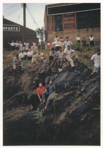 photograph of a group of people sitting on a rocky hillside near some industrial buildings all looking at the camera