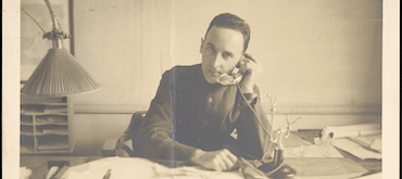 Sepia-toned historical photograph of a man in a military uniform sitting at a desk and using a vintage candlestick telephone.