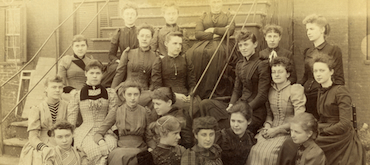 Sepia-toned historical photograph of a large group of young women in high-collared Victorian-era dresses, posed on the steps outside a brick building.