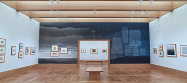 A gallery with light wood floors and a large dark blue-and-grey painted mural along the back wall; framed works hang on the white side walls, with a low wooden bench at center.