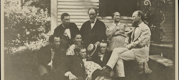 A sepia-toned historical photograph of a group of men and women dressed in 19th-century attire, gathering outdoors next to the porch steps of a clapboard house.