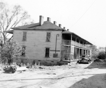 A two-story boarding house situated on a dirt road, with stacked front porches and broken windows.