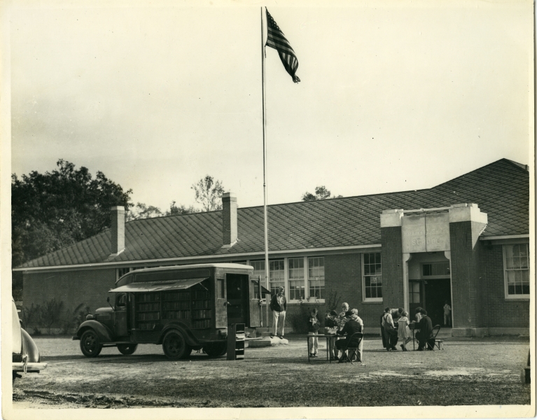 HCPL Bookmobile parked in front of the Smiley School.  Two librarians sit at tables and help students borrow books.