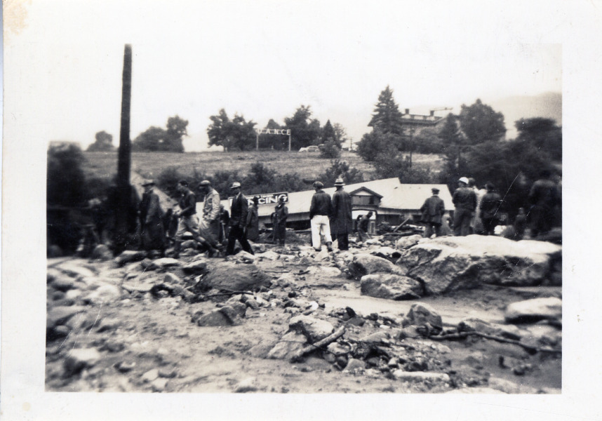 Group of men surveying the damage in the aftermath of a flood of Bear Creek on the town of Morrison. Taken from in front of the garage facing Dance Hall, the men all standing on top of damaged surface facing the Hall.