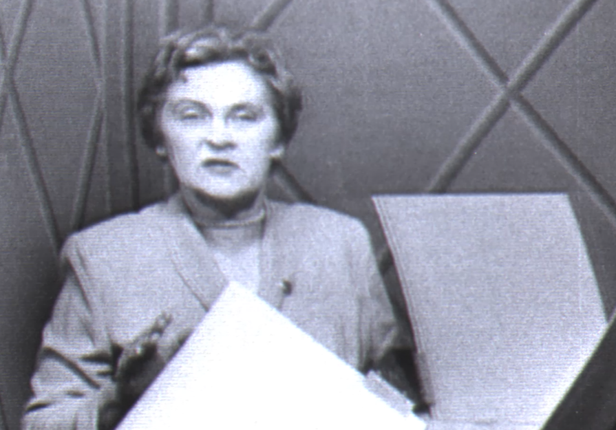 A black and white film still of a woman speaking directly to the camera. A solar device is in the foreground.