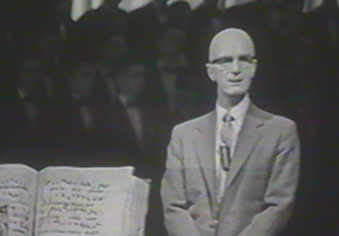 A black and white still frame of a man, program host Lynn Poole, speaking to the camera, with a large book in the lower left of the frame.