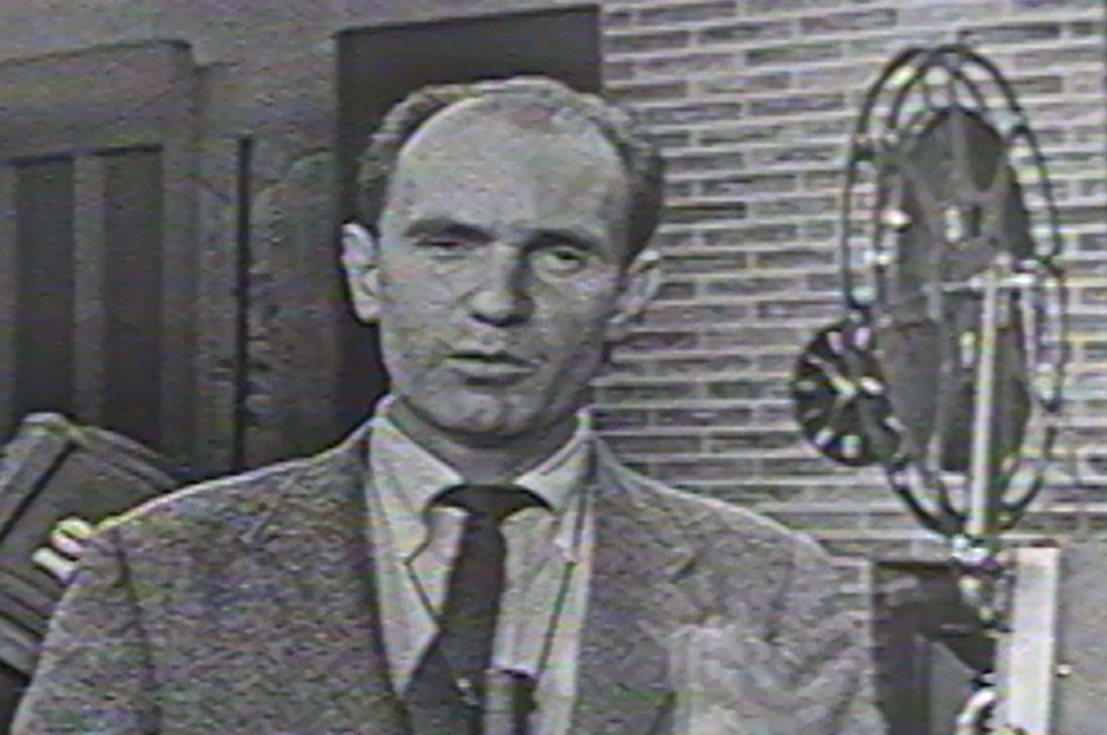 A black and white still frame of a man speaking to the camera. To the right of him is a reel-to-reel film projector.