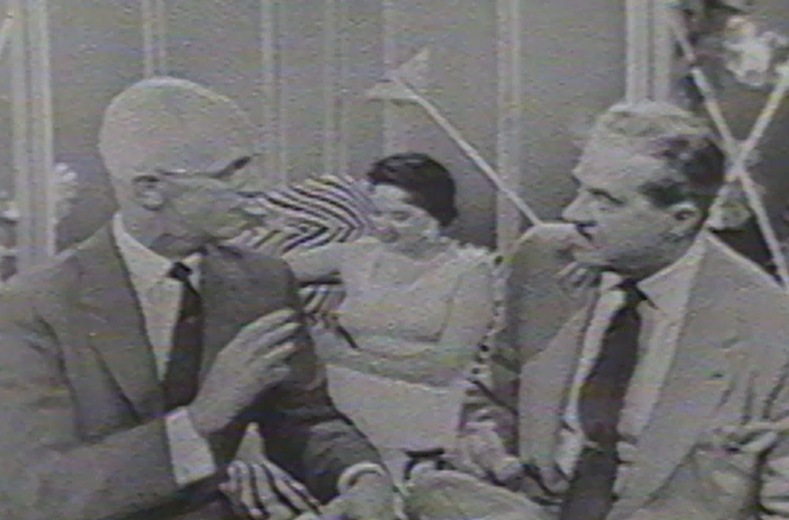 A black and white still frame of two men talking, while a woman reads in the background.