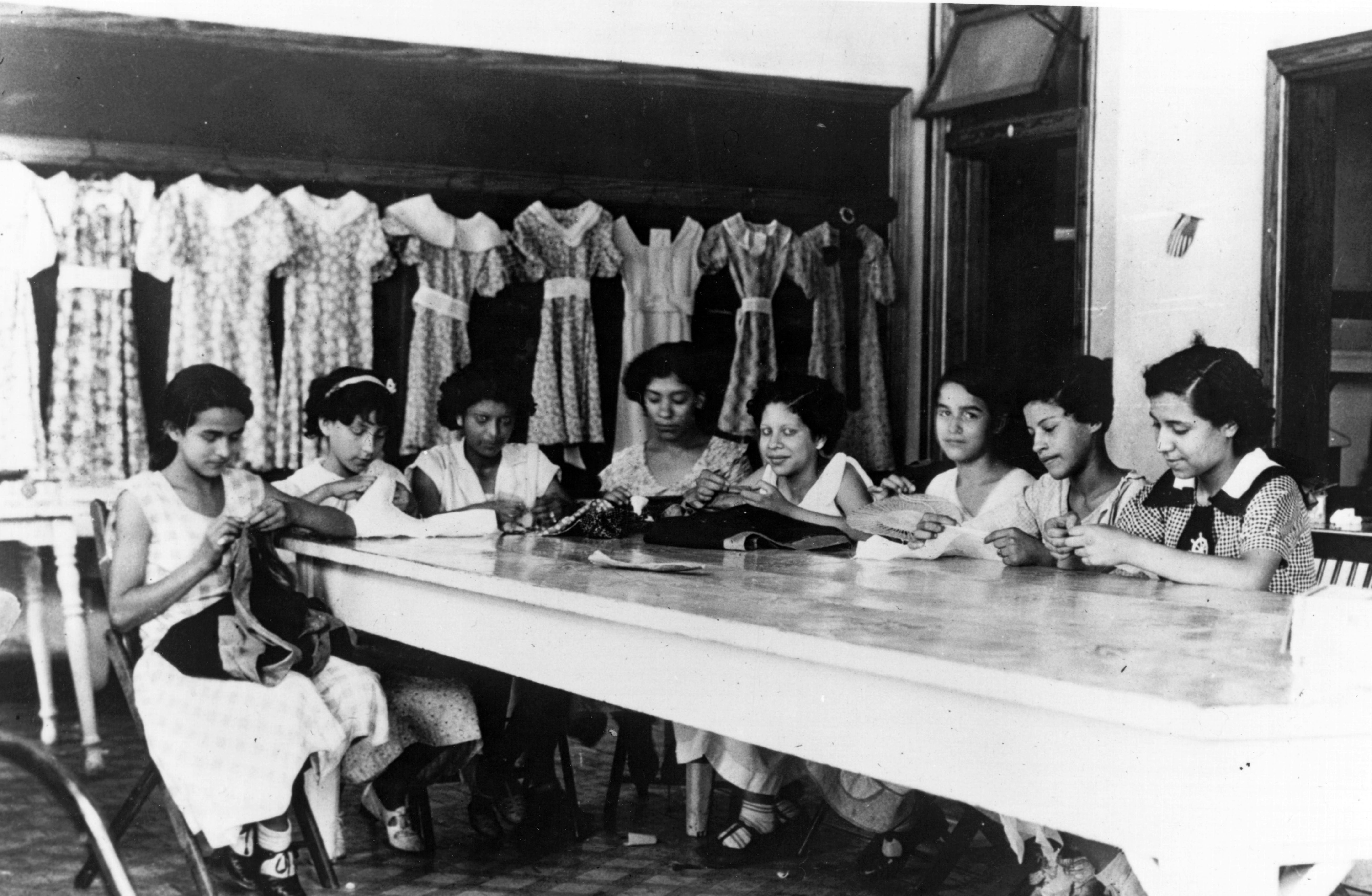 Eight women seated at a tall table sewing garments