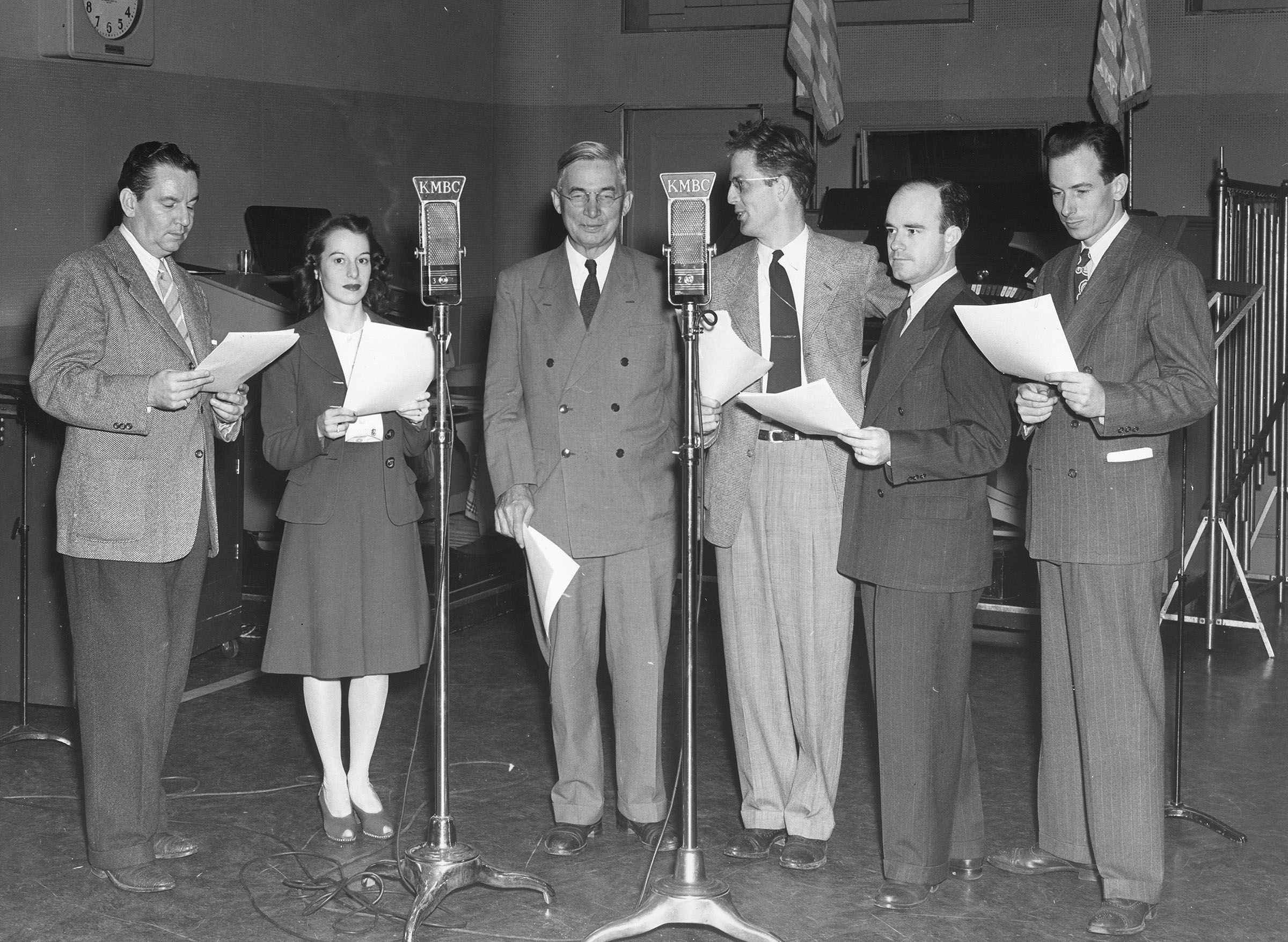 Five men and one woman standing behind two microphones on tall stands