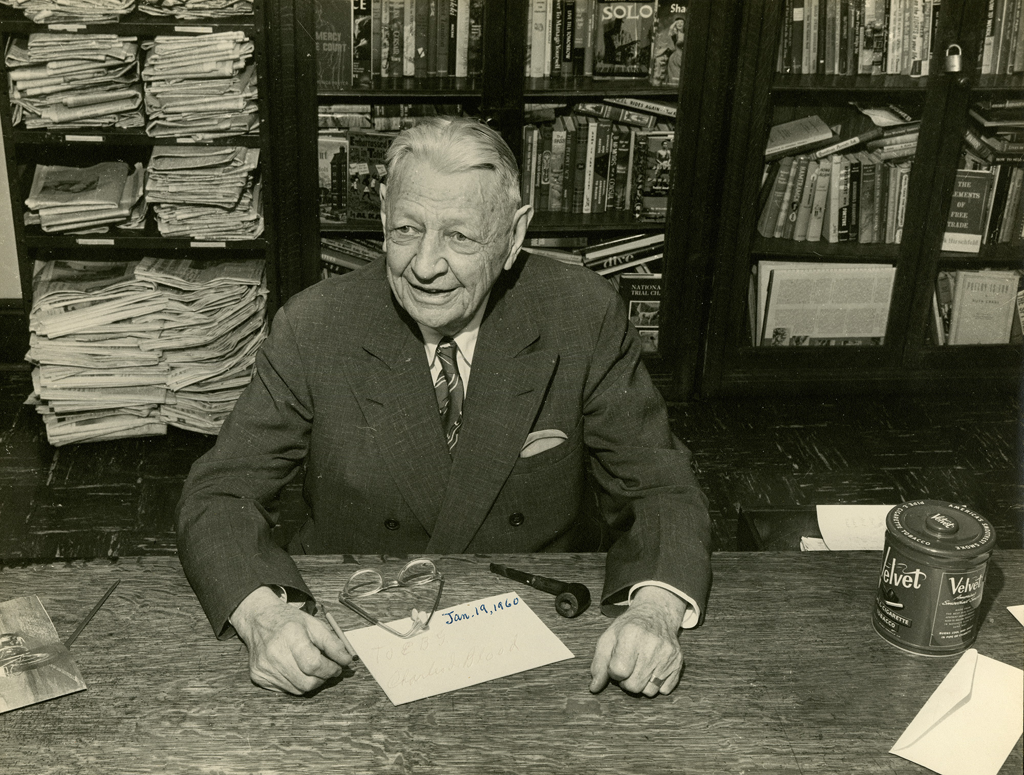 An older man seated in front of a shelf full of papers and books.