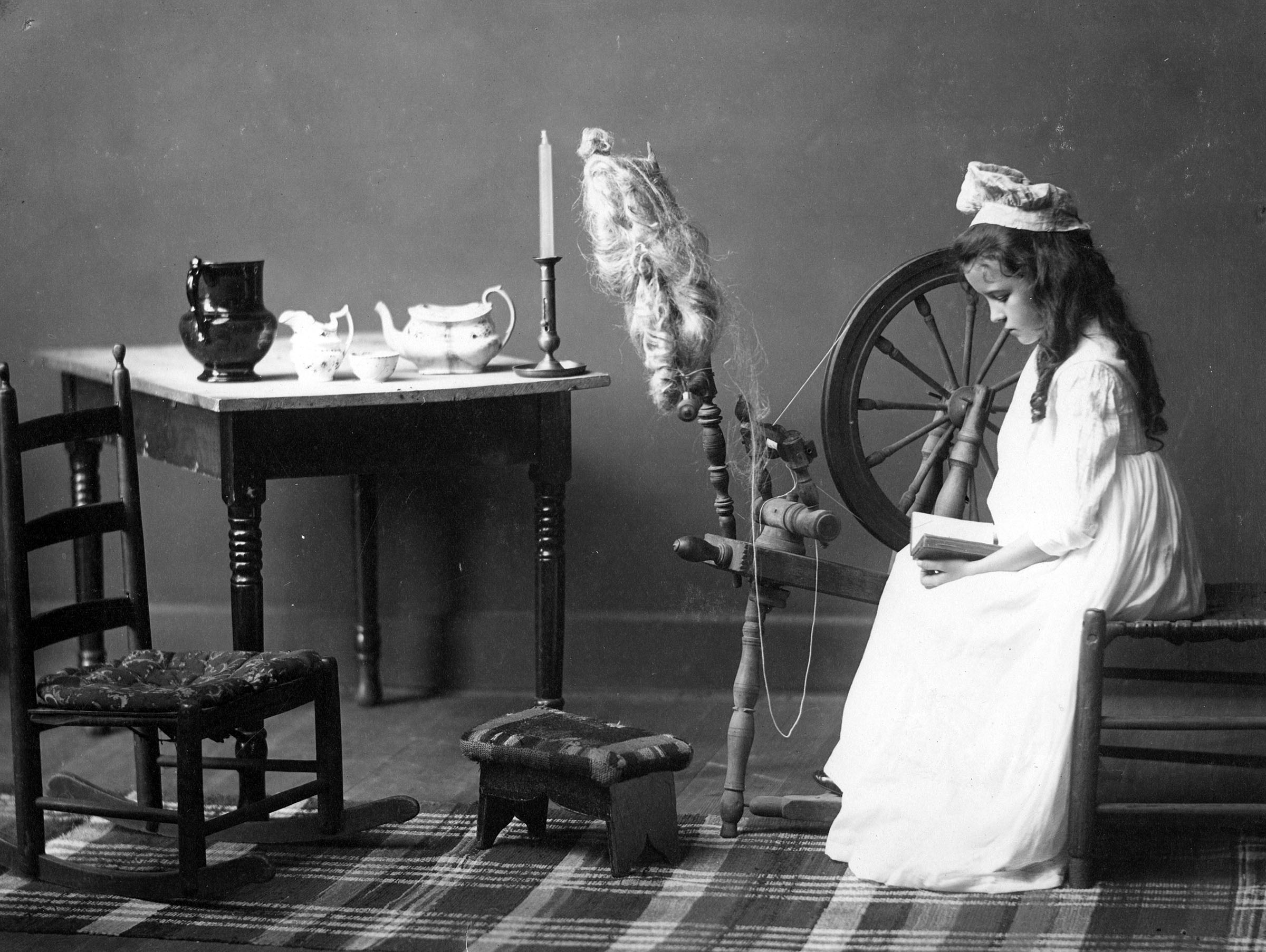 A young girl with a white dress and bonnet sitting at a spinning wheel