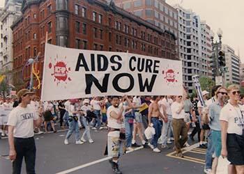 A demonstration march on a city street. A banner reading "AIDS Cure Now" is featured front and center.