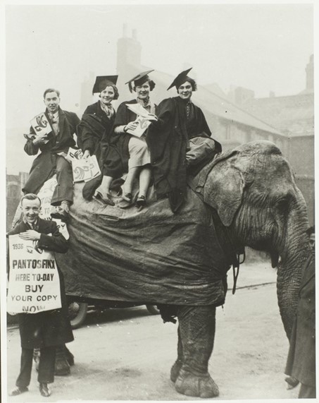 Students in mortar boards and gowns sitting on an elephant with placards to advertise a student magazine