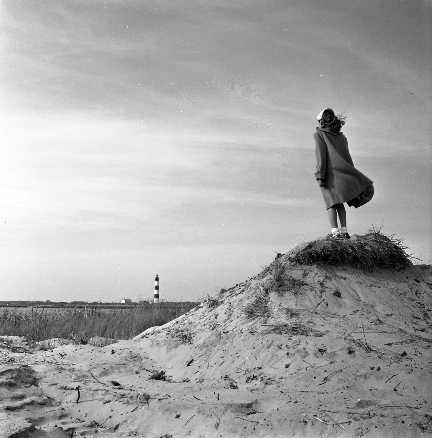 Photo of a girl or young woman standing on a sand dune looking toward a lighthouse