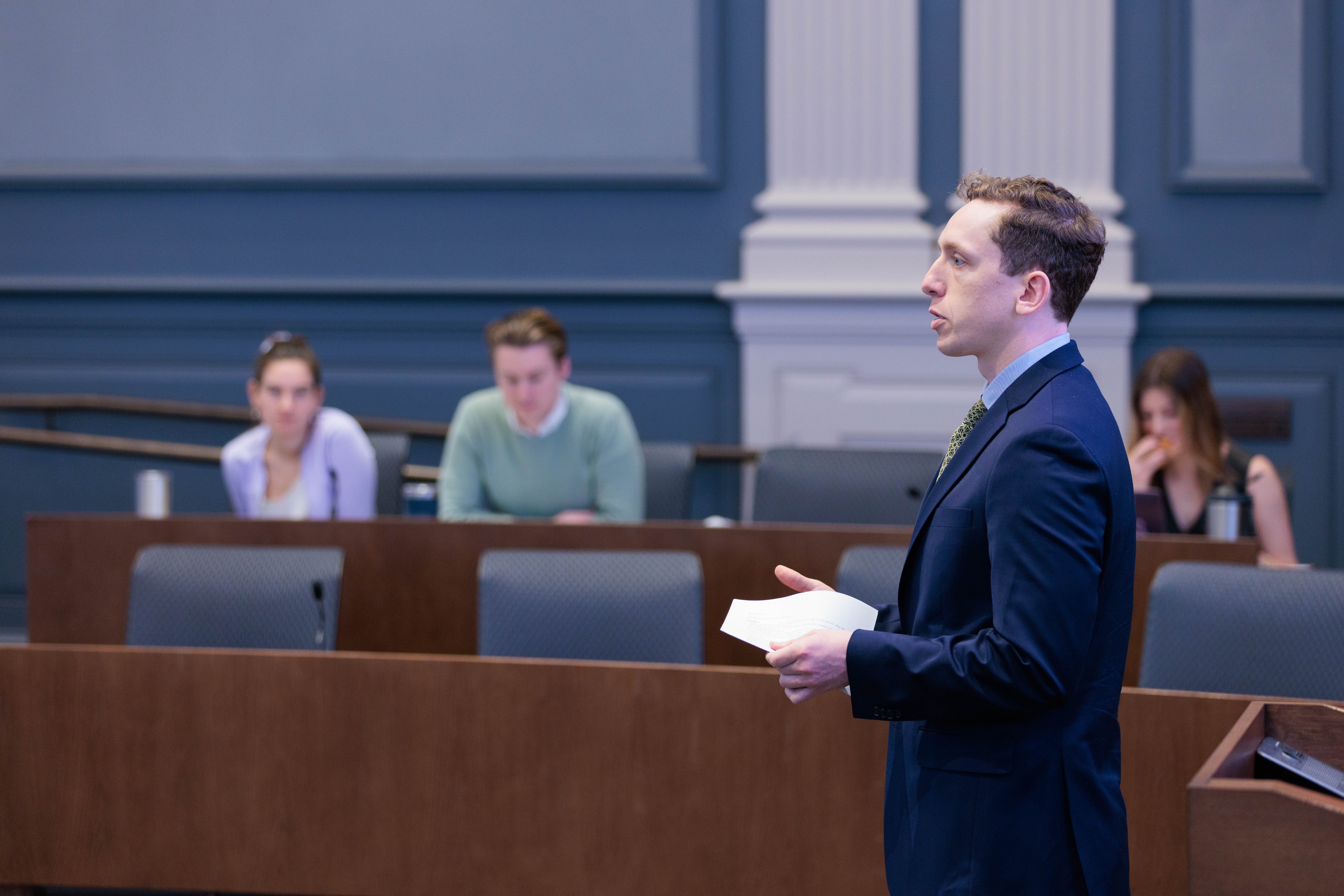 A photo of a white male students in a blue jacket addressing the room.