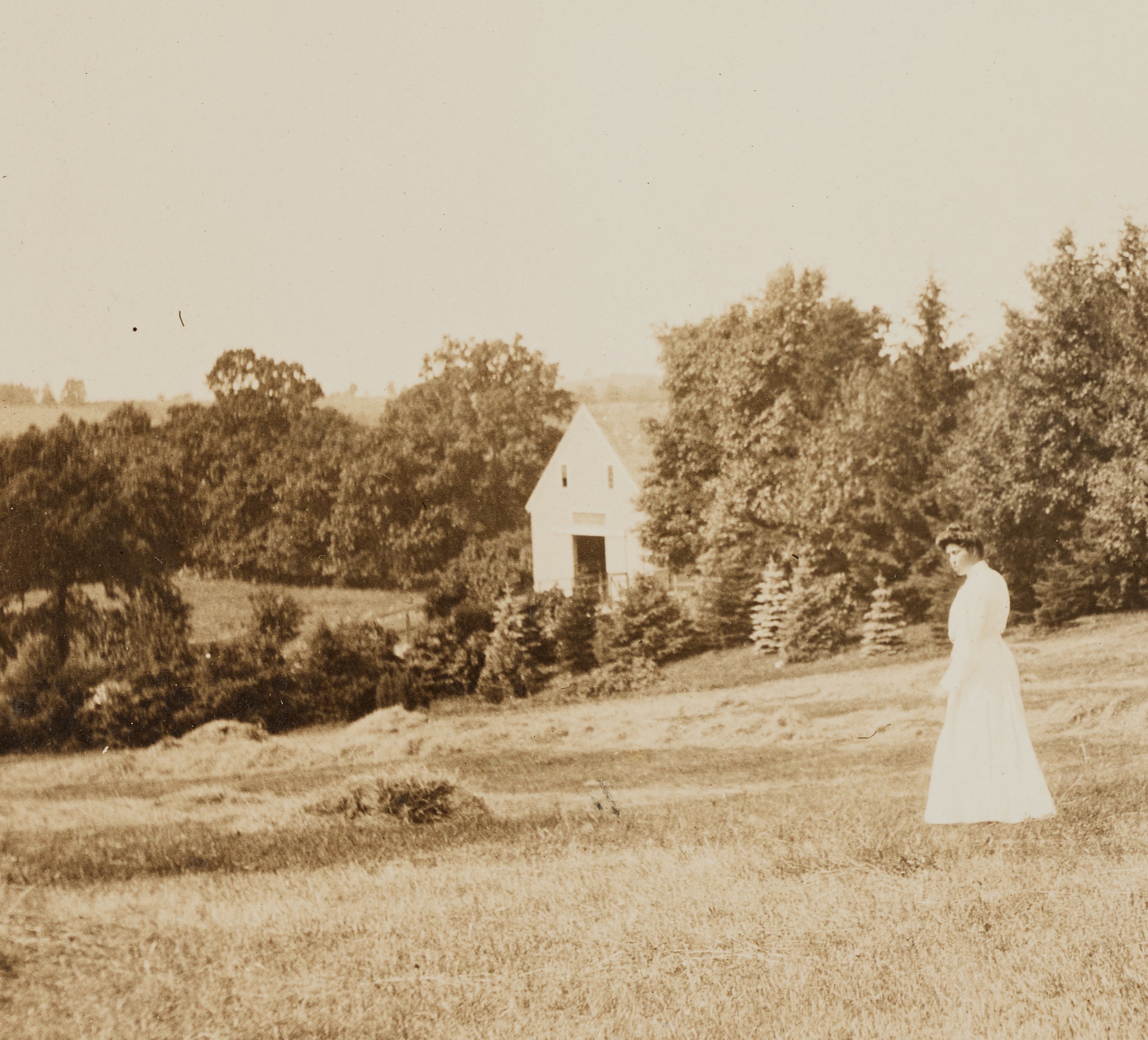 Outdoor scene depicting a field of grass with trees and a white house in the background. A women in a white dress on the right side of the frame is walking in the foreground.