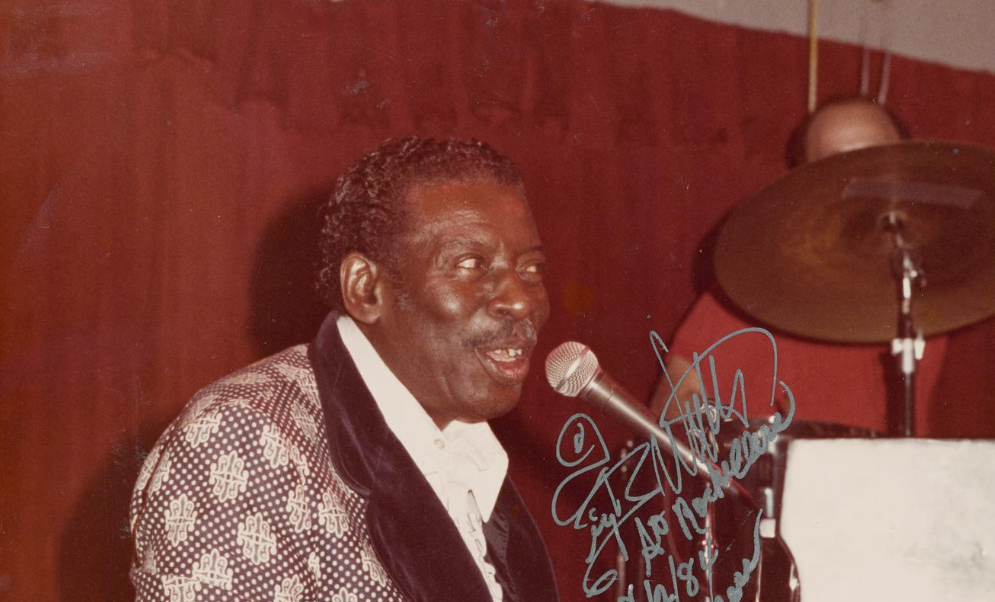 Big Walter Price wearing an ornate jacket that is maroon with lace detailing and a velvet lapel with a white dress shirt. He's singing into a microphone and playing a piano in front of a wood paneled wall.