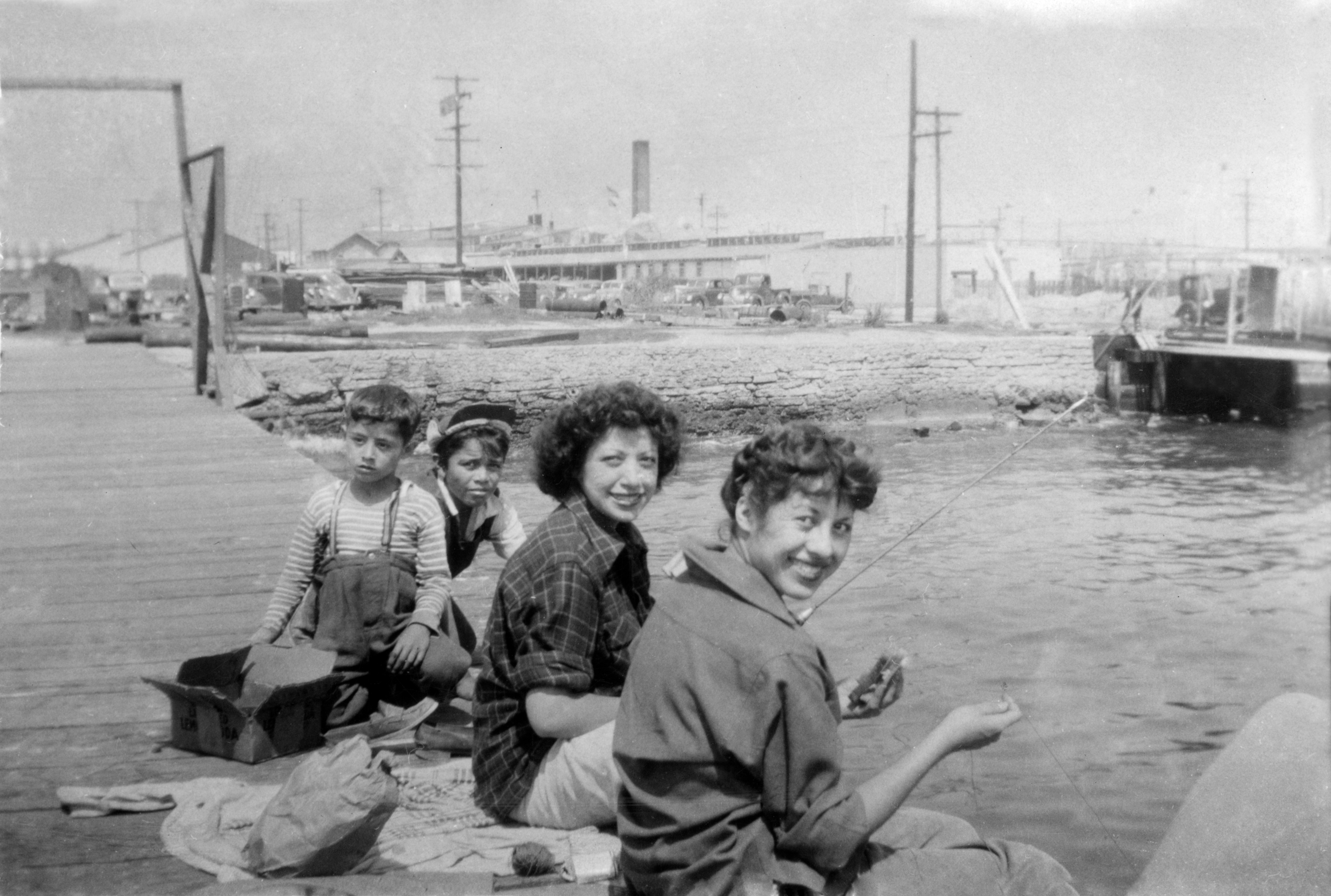 A black and white photo of three women and a young child sitting on the dock of a marina with boats in the background.