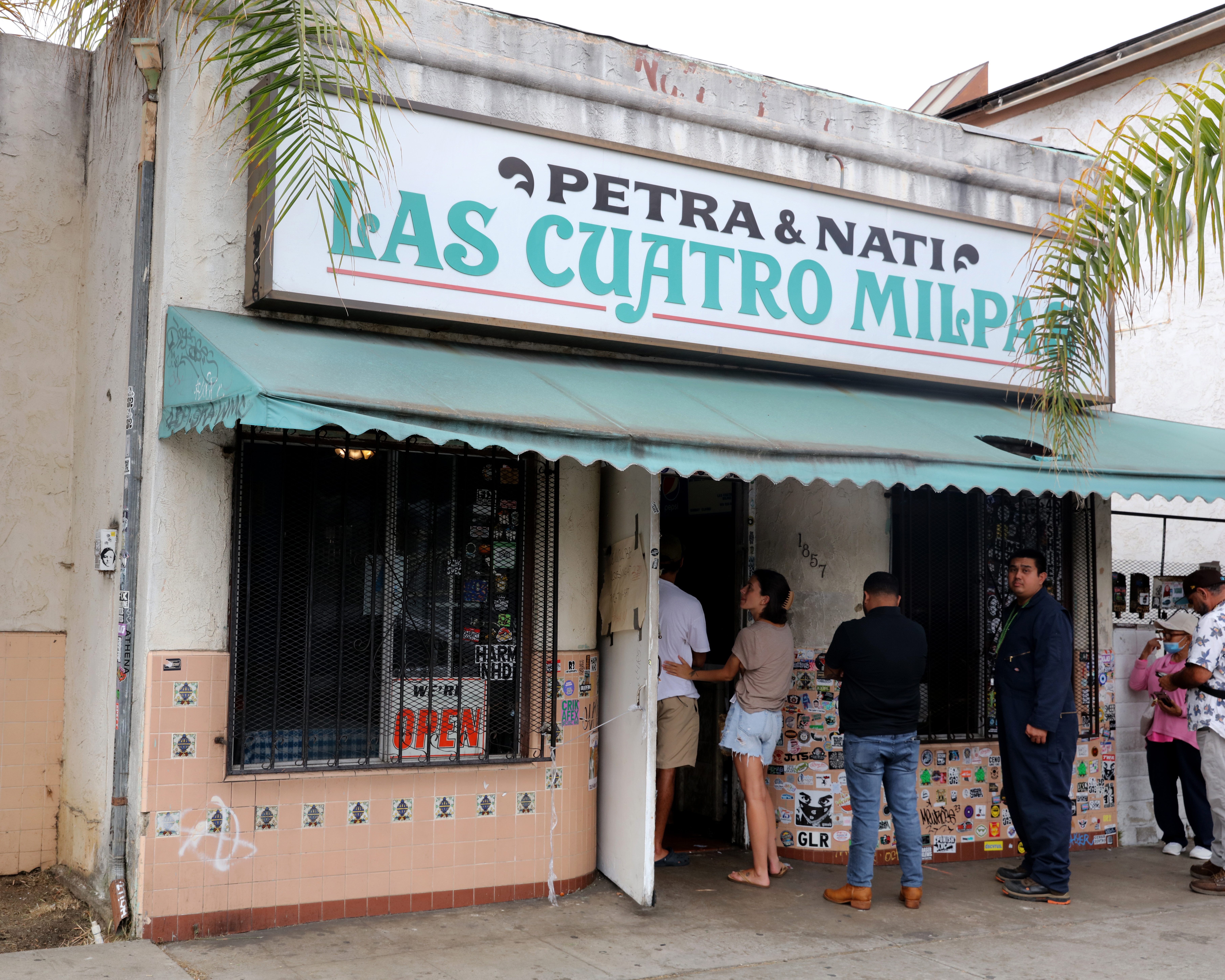 A color image of people standing in line to enter a building. Above the door is a sign and green awning. The sign reads "Petra and Nati Las Cuatro Milpas."
