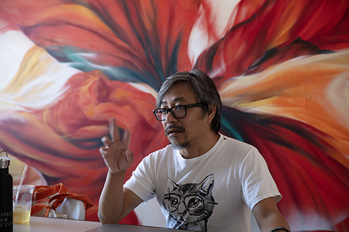 A color photo of an middle-aged asian man sitting in front of a wall with a floral pattern.