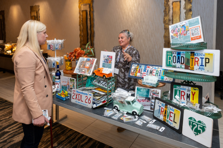 A picture of a woman at a table holding artwork made out of license plates talking to another woman.