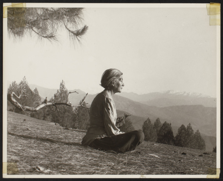 A photo of a woman sitting crossed legged, eyes closed, facing the right, down a hill with mountains in the background.