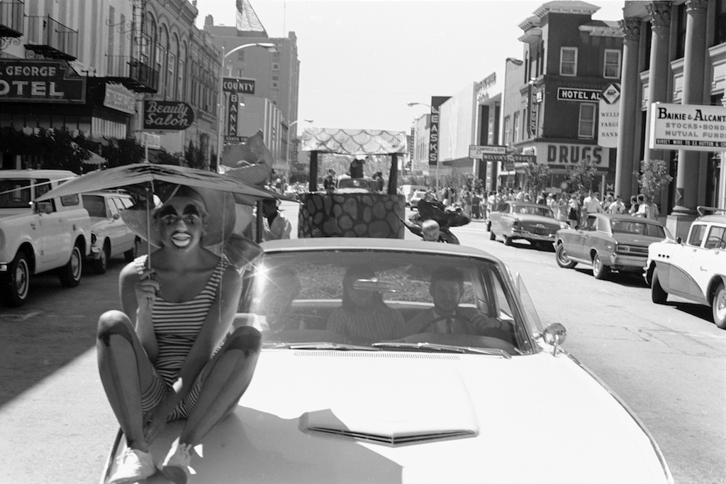 B&W photo of Spring Parade, person in clown makeup sitting on the hood of a car in the street
