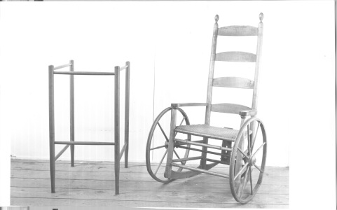 Antique black-and-white image of a square shaped wooden walker, alongside a wooden wheelchair created from a recycled rocking chair mounted on two wooden wheels.