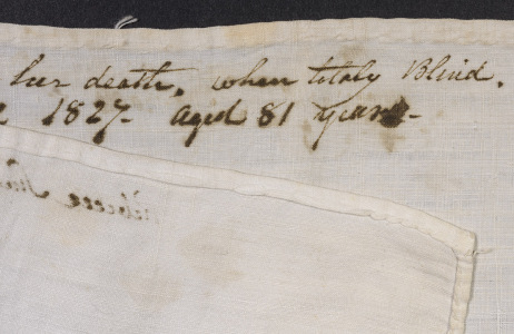 White handkerchief with an ink inscription with one corner folded over and across itself, revealing the stitches of the hem.
